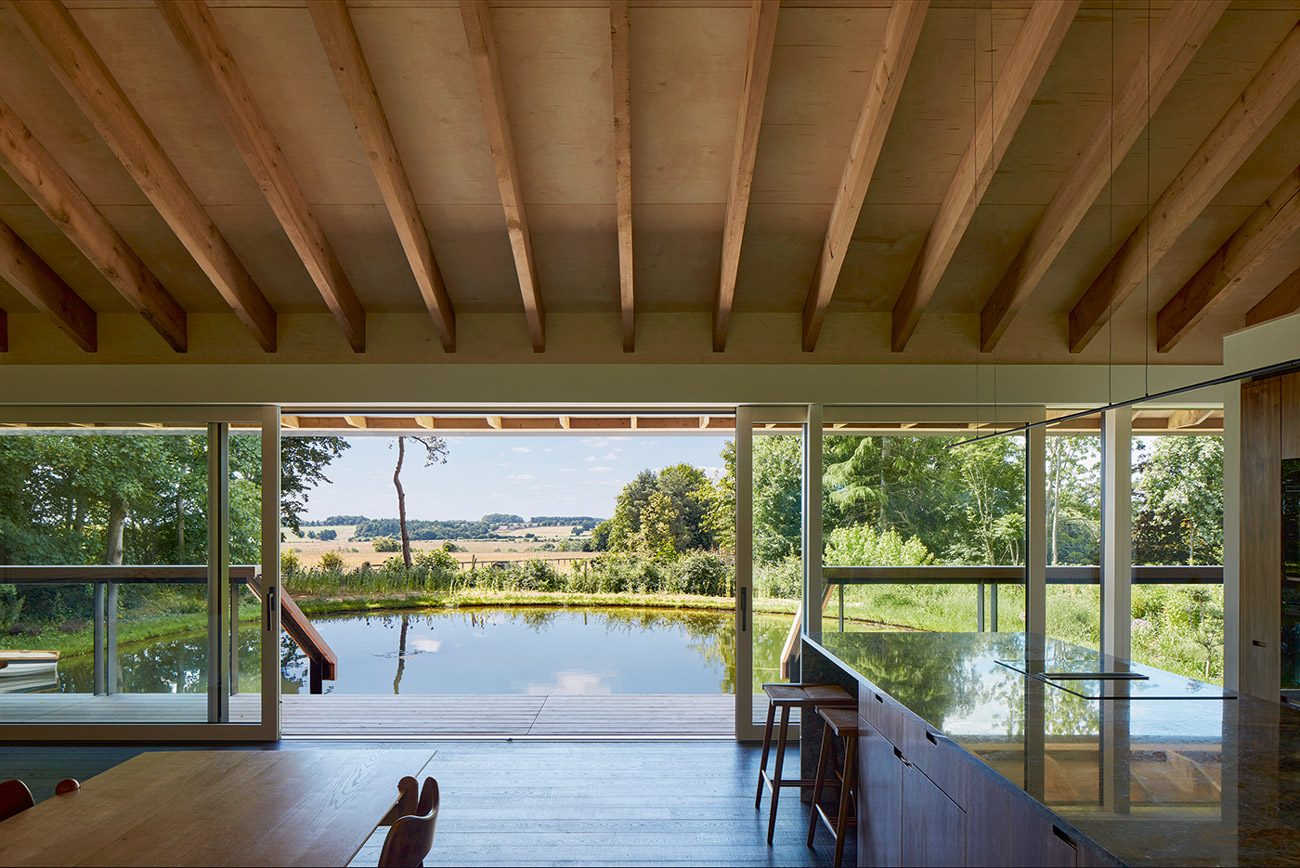 The living space with the kitchen to the right and four-panel picture windows to the pond and fields beyond.